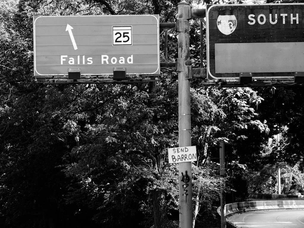 Overhead Falls Road and SOUTH signs above a signpost with a small SEND BARRON sign