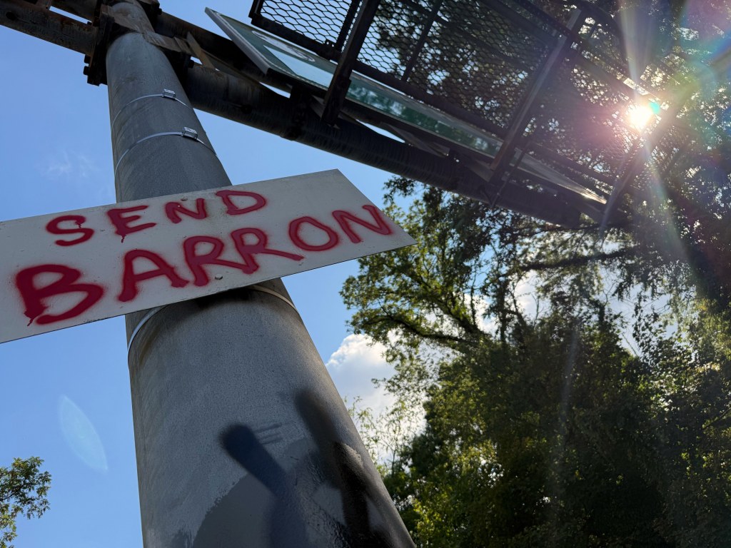 SEND BARRON sign seen from below with the sun behind the overhead signs